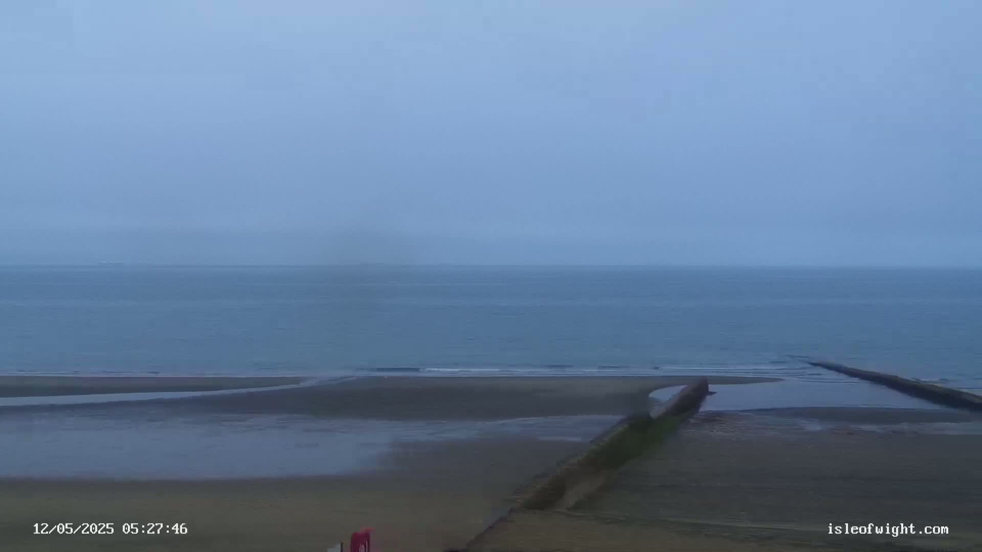 A calm, overcast day shows a beach with a low tide, a dark wall-like structure extending into the water, and a flat, gray sea.