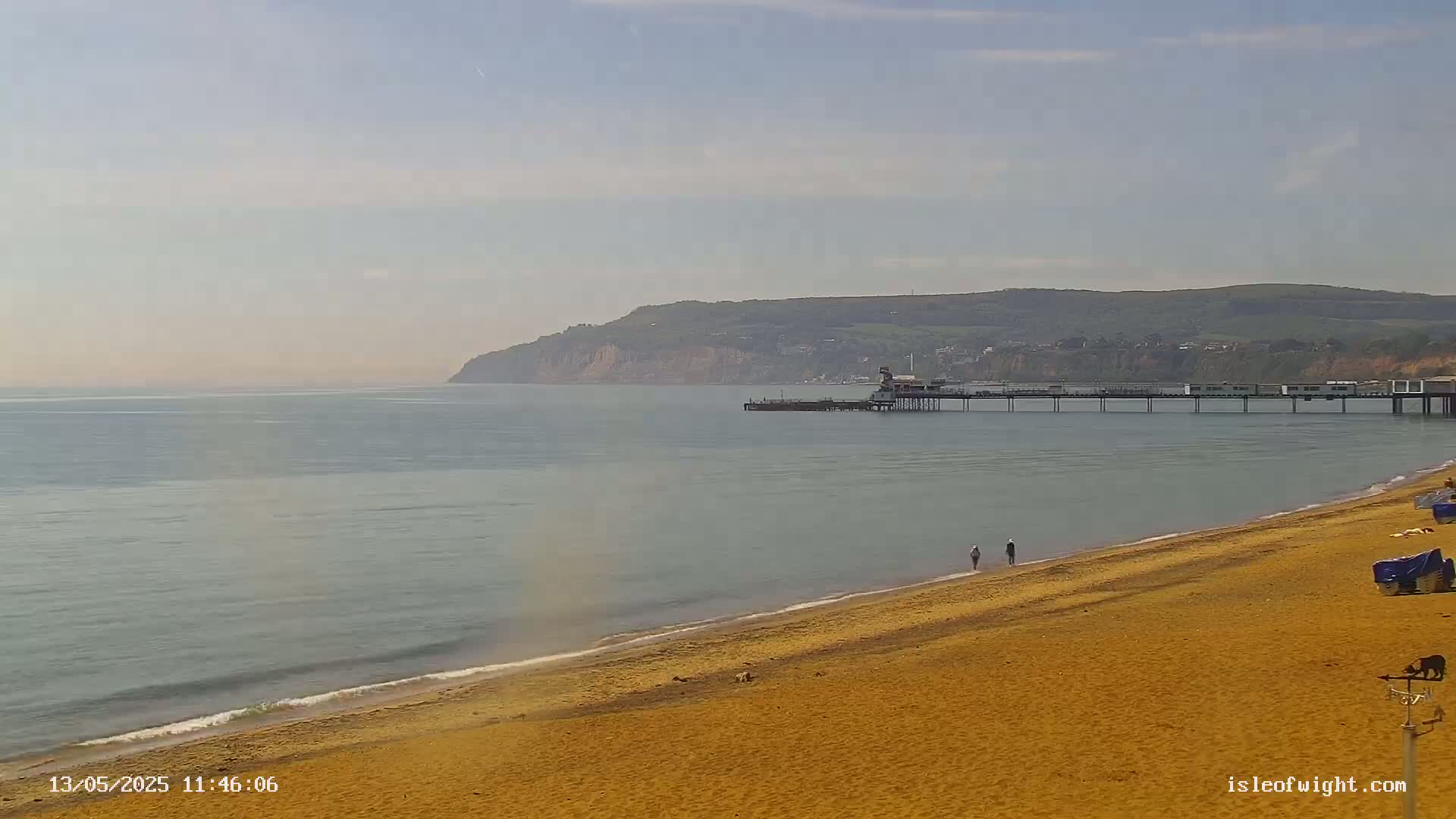 A sandy beach stretches toward a calm sea and a pier extending to a distant, hilly coastline under a hazy sky.