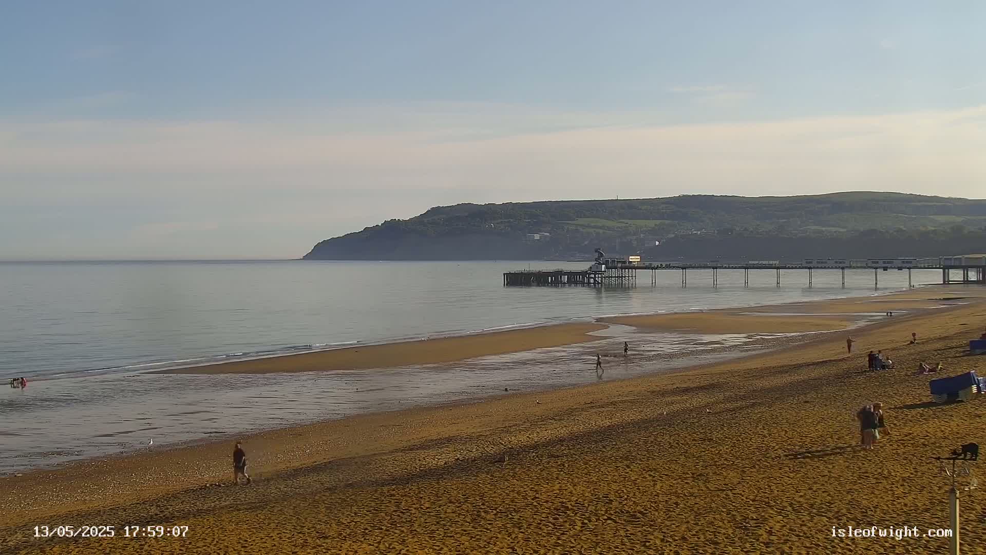 A long sandy beach stretches out to a pier extending into a calm sea, with a hill visible in the background under a clear sky.