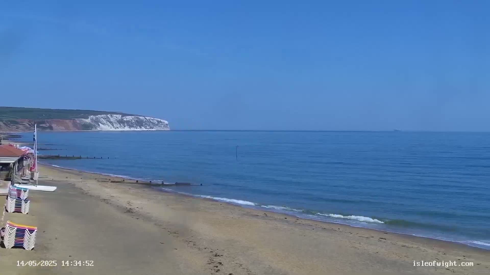 A sandy beach with calm ocean waves stretches toward a distant white cliff face under a clear blue sky.