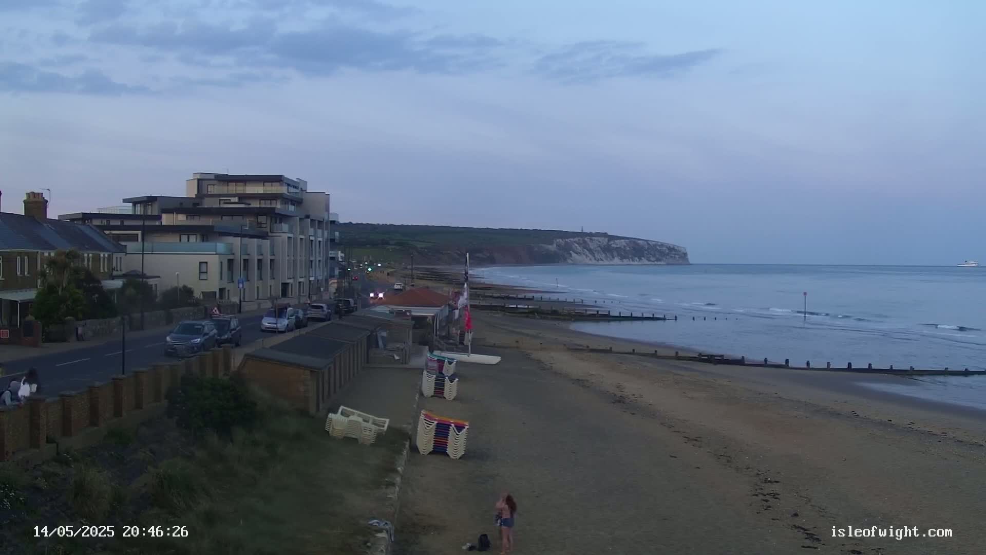 A sandy beach with stacked beach chairs, a few people, and a modern building is seen near a calm ocean under a twilight sky, with a large cliff visible in the distance.