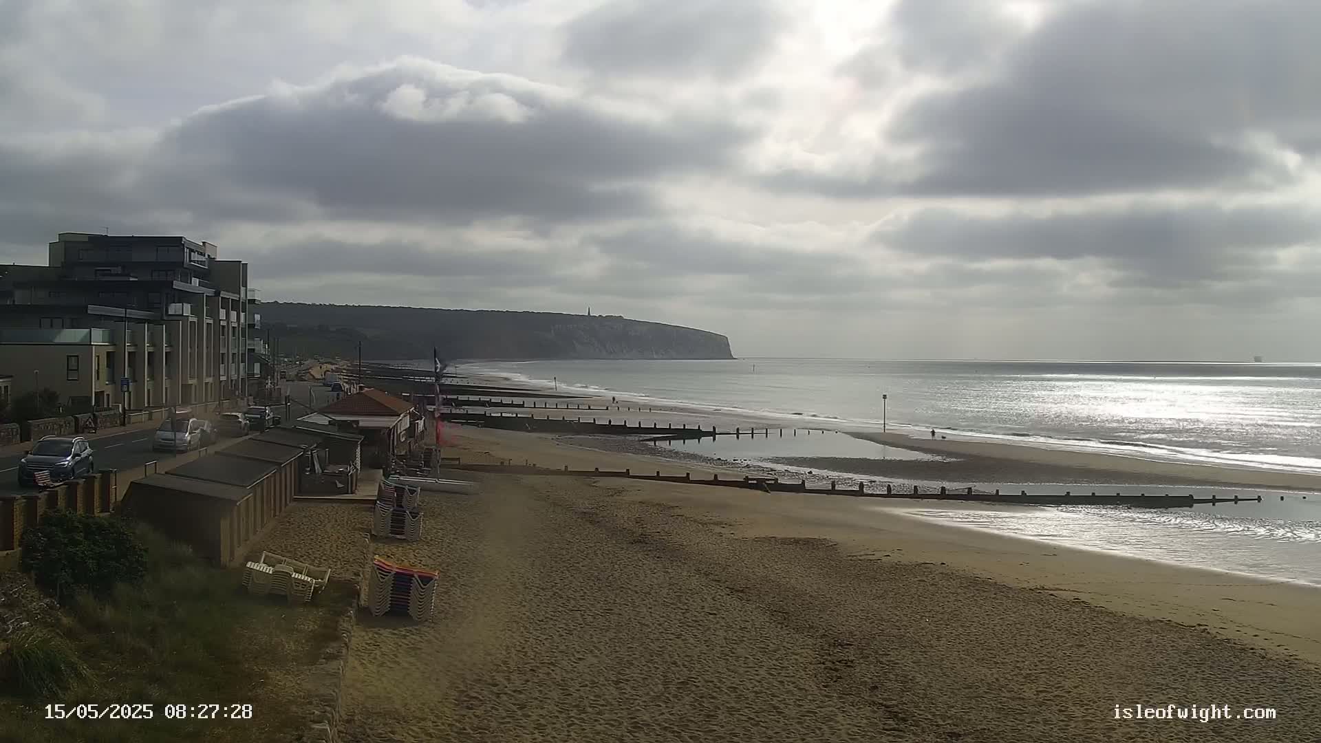 A sandy beach stretches out to a calm sea under a cloudy sky, with a building and beach huts visible on the left.