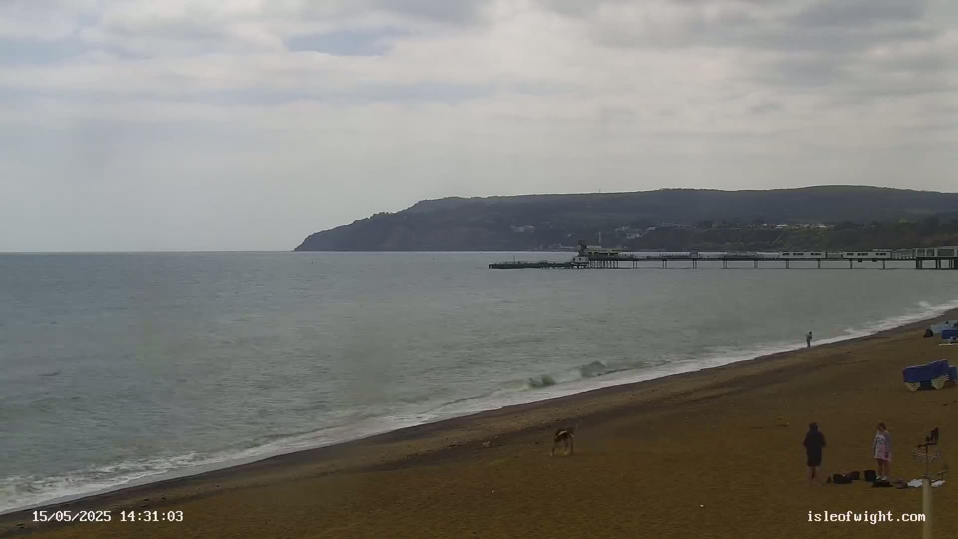 A sandy beach with a few people, a pier extending into a calm sea, and a distant hill under an overcast sky.