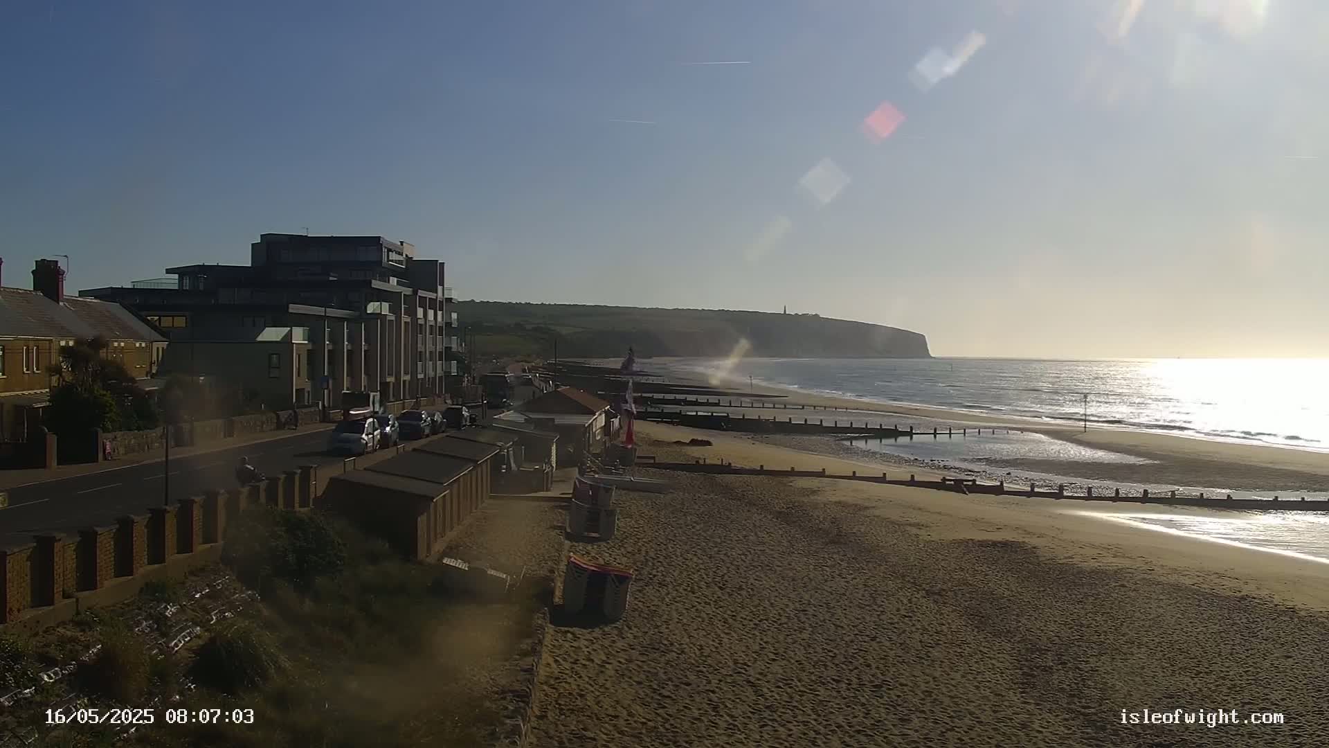 A sandy beach with calm ocean waves stretches out before a row of beach huts and buildings, under a clear, sunny sky.