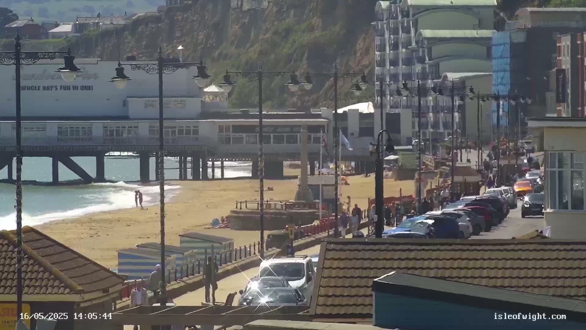 A sunny day at a beach town shows a pier, sandy beach with people, a promenade with parked cars and pedestrians, and buildings on a cliff overlooking the scene.