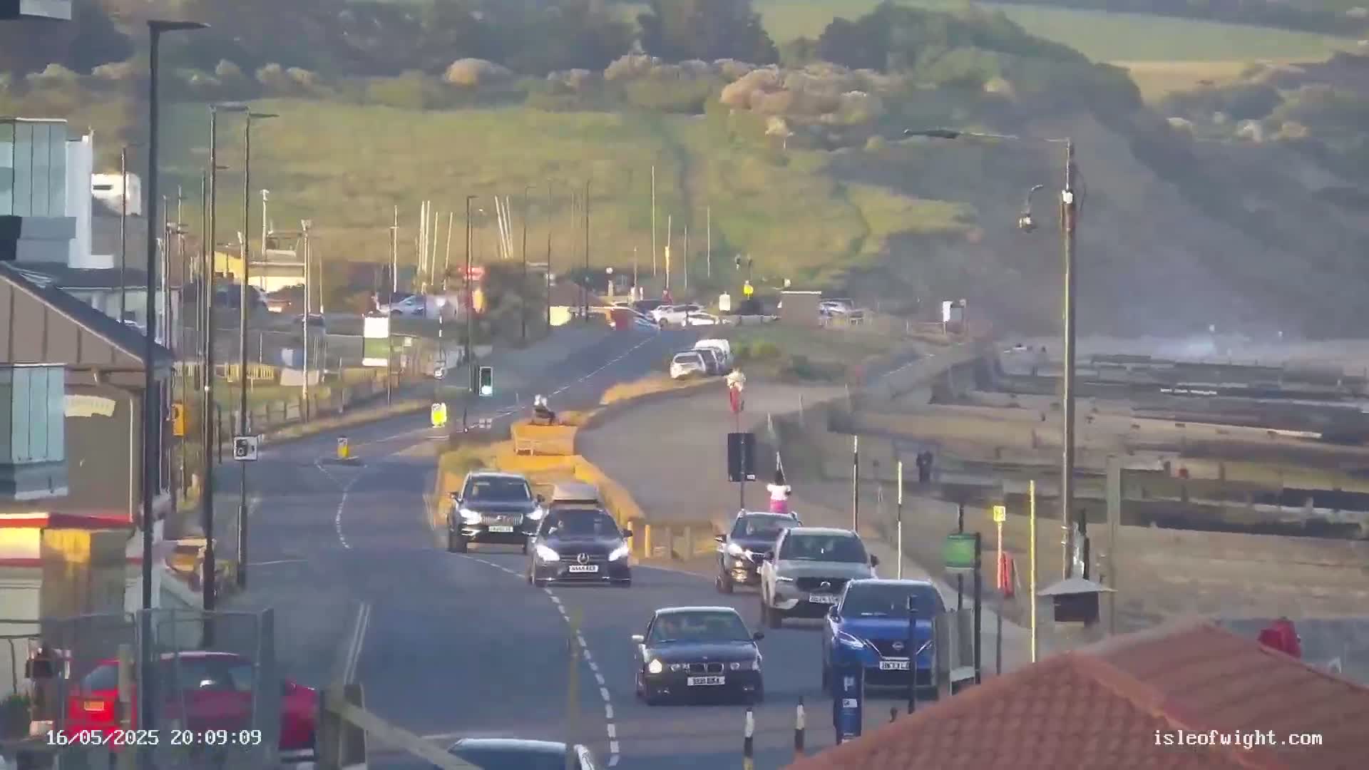 Several cars drive along a coastal road under a partly sunny sky, with choppy waves visible in the background.