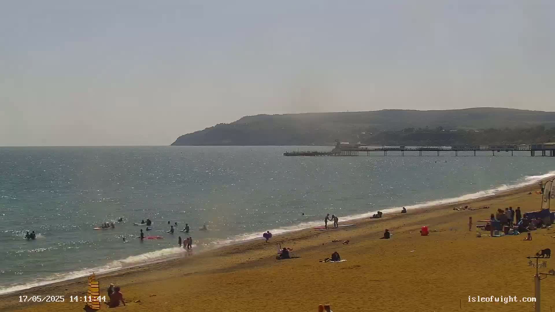 A sandy beach with people swimming and sunbathing, a pier extending into a calm sea under a clear sky, and hills visible in the distance.