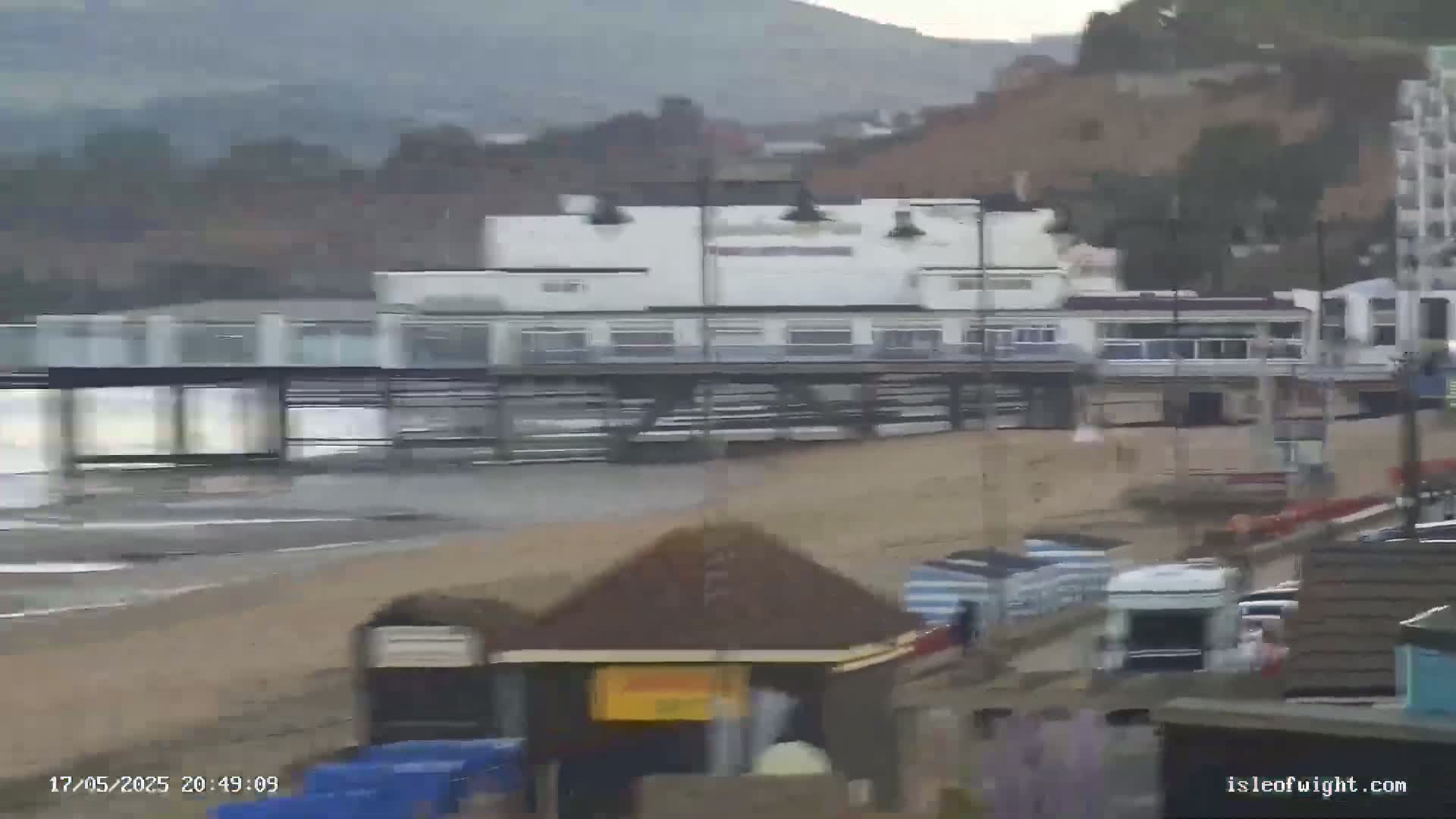 A long pier with a building on it sits on a sandy beach, with a somewhat overcast sky and choppy water.