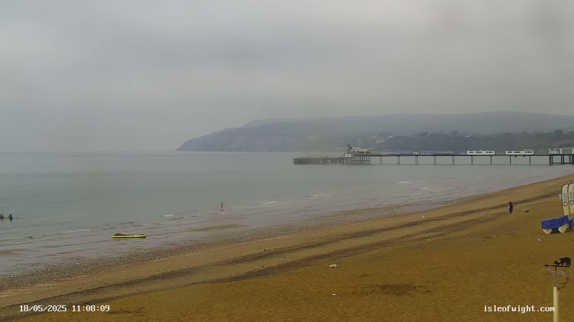 A long pier extends into a calm, grey sea under an overcast sky, overlooking a sandy beach with a few people and a small inflatable boat.