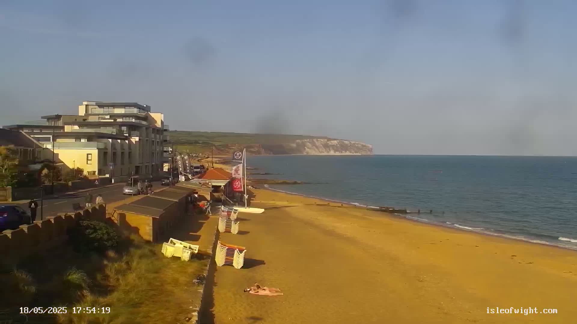 A sandy beach stretches below a multi-story building near calm ocean water under a clear sky, with white cliffs visible in the distance.