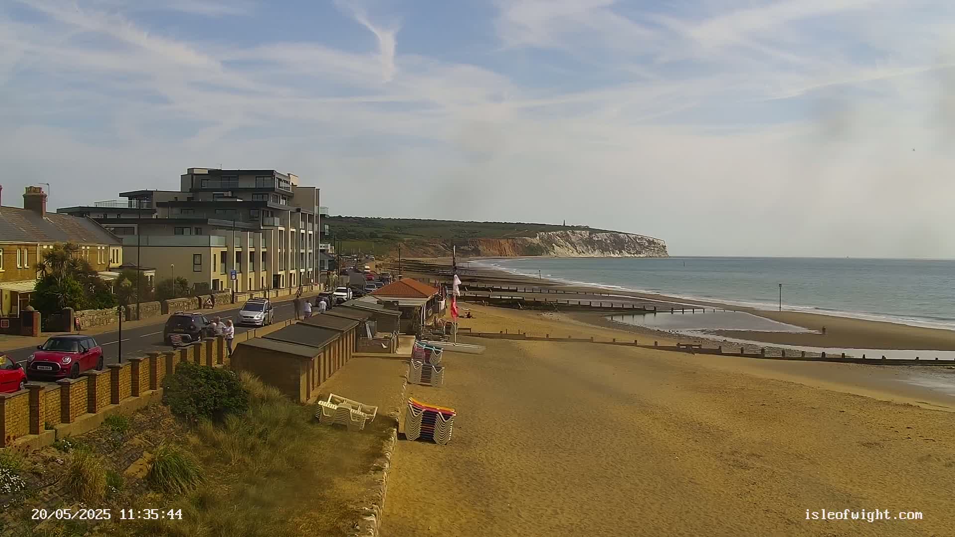 A sandy beach with calm ocean waves stretches alongside a road lined with buildings, beach huts, and people, under a partly cloudy sky.