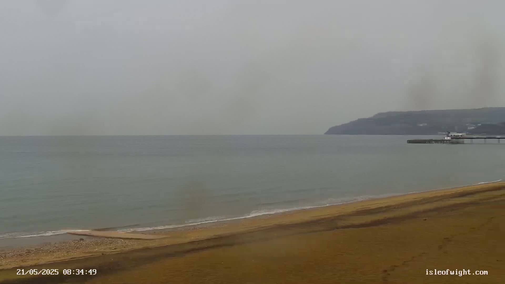 A sandy beach meets a calm, gray sea under an overcast sky, with a pier and distant cliffs visible in the background.