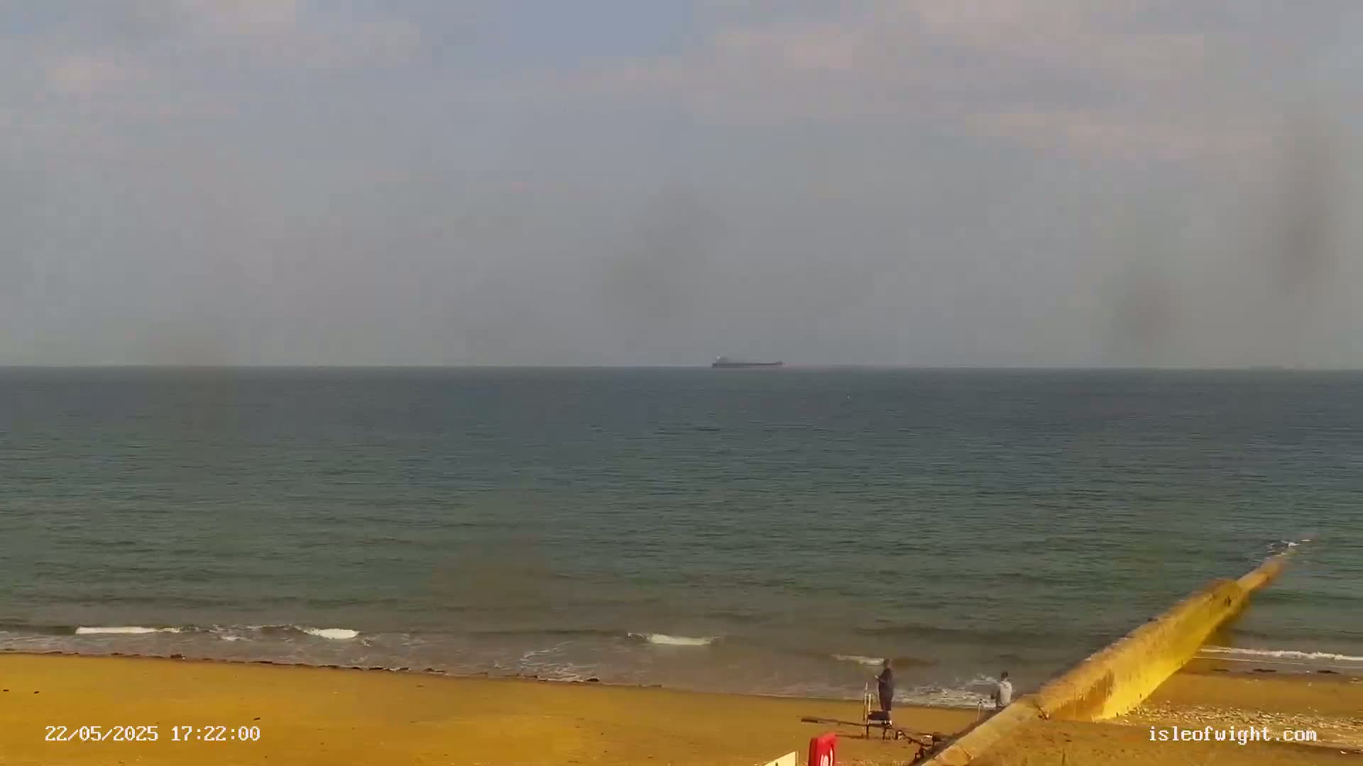 A calm, overcast day at a beach shows a distant cargo ship on the horizon, with a few people standing near a low seawall on a sandy shore.