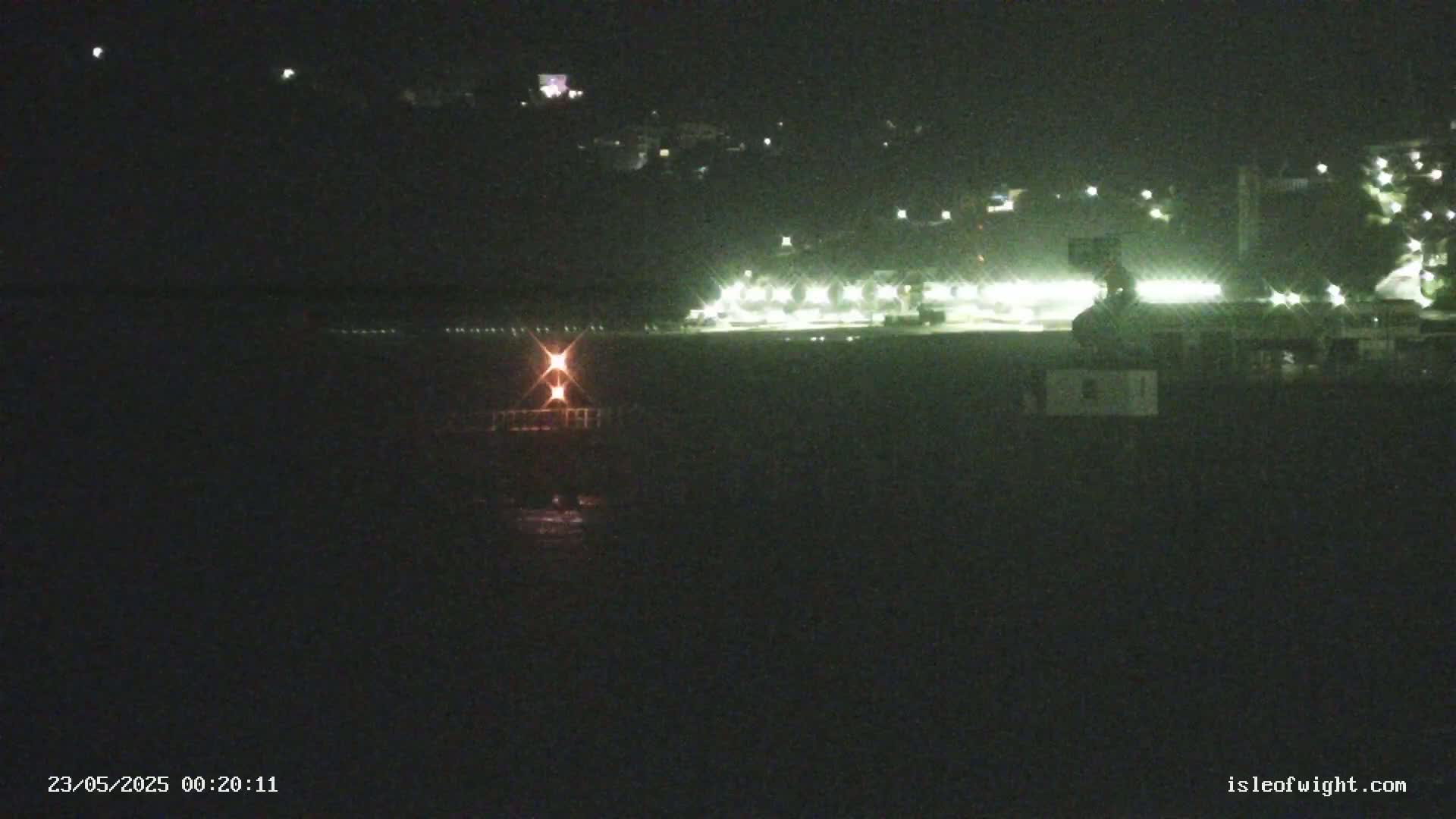 A nighttime long shot shows a body of dark water in front of a brightly lit waterfront area with buildings and lights on a hill in the background.