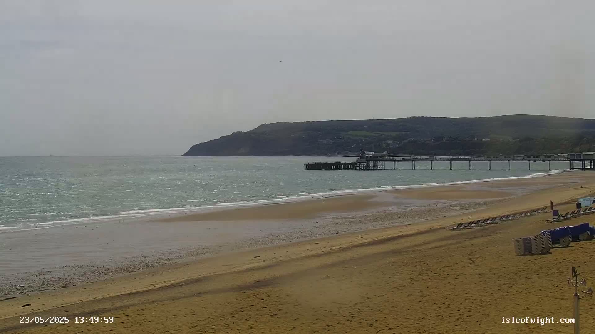 A sandy beach with a pier extending into a calm, light-green sea under an overcast sky, with a hill visible in the background.