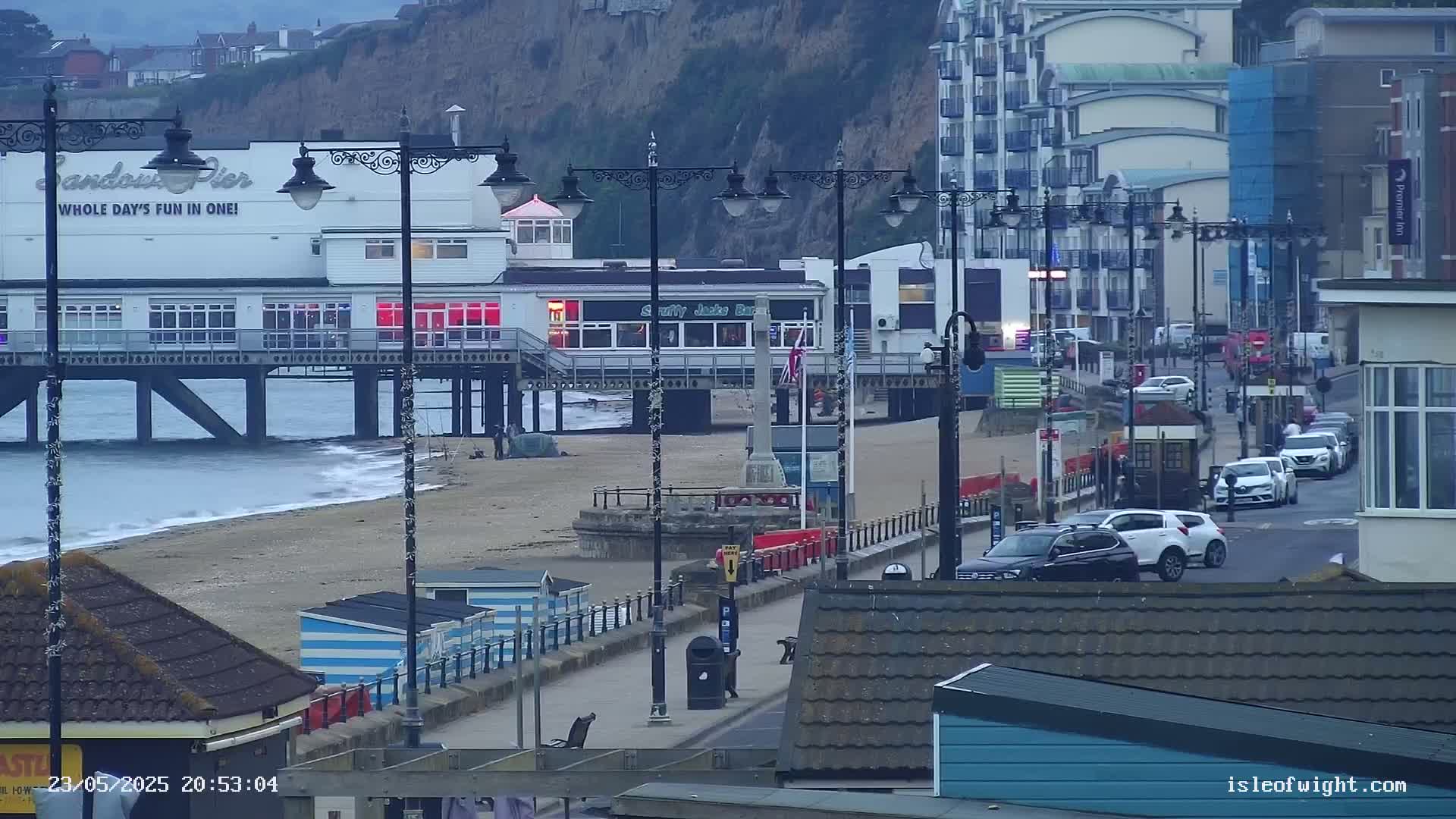 A pier with shops and restaurants extends over a sandy beach, alongside a road with parked cars, all under an overcast sky.