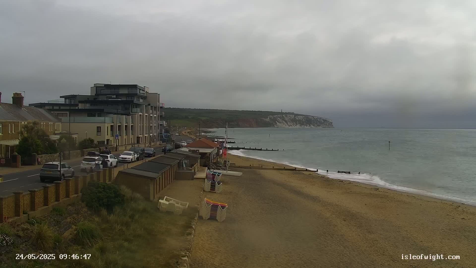 A sandy beach with parked cars and beach huts stretches along a calm sea under an overcast sky, with a multi-story building and cliffs in the distance.