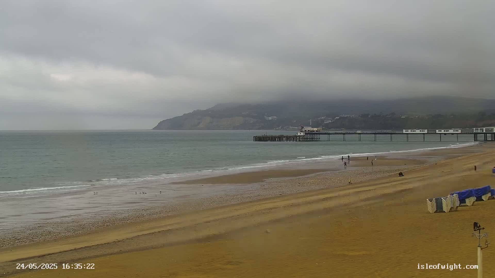 A long pier extends from a sandy beach into a calm sea under an overcast sky, with a distant hill visible in the background.