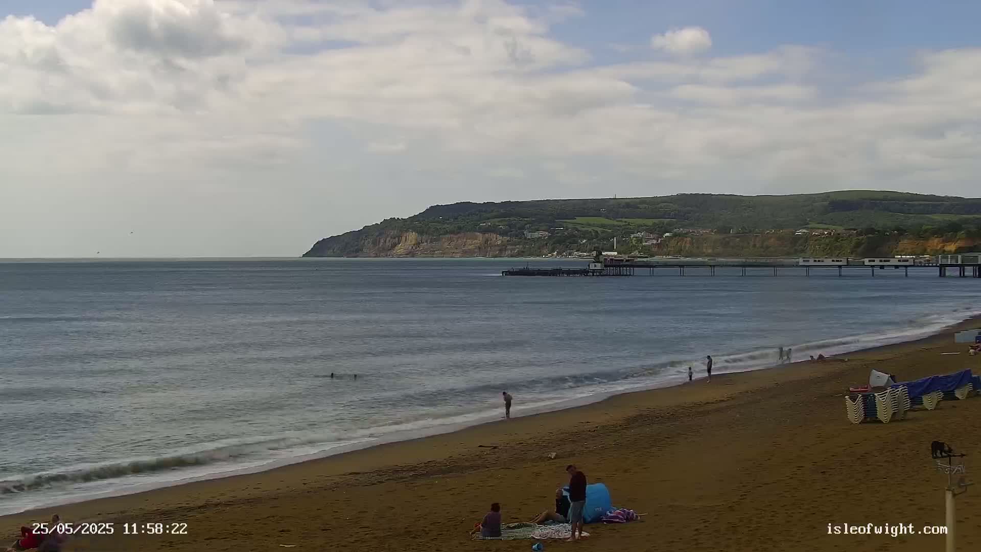 A sandy beach with a few people on it stretches toward a pier and a large cliff in the distance under a partly cloudy sky.