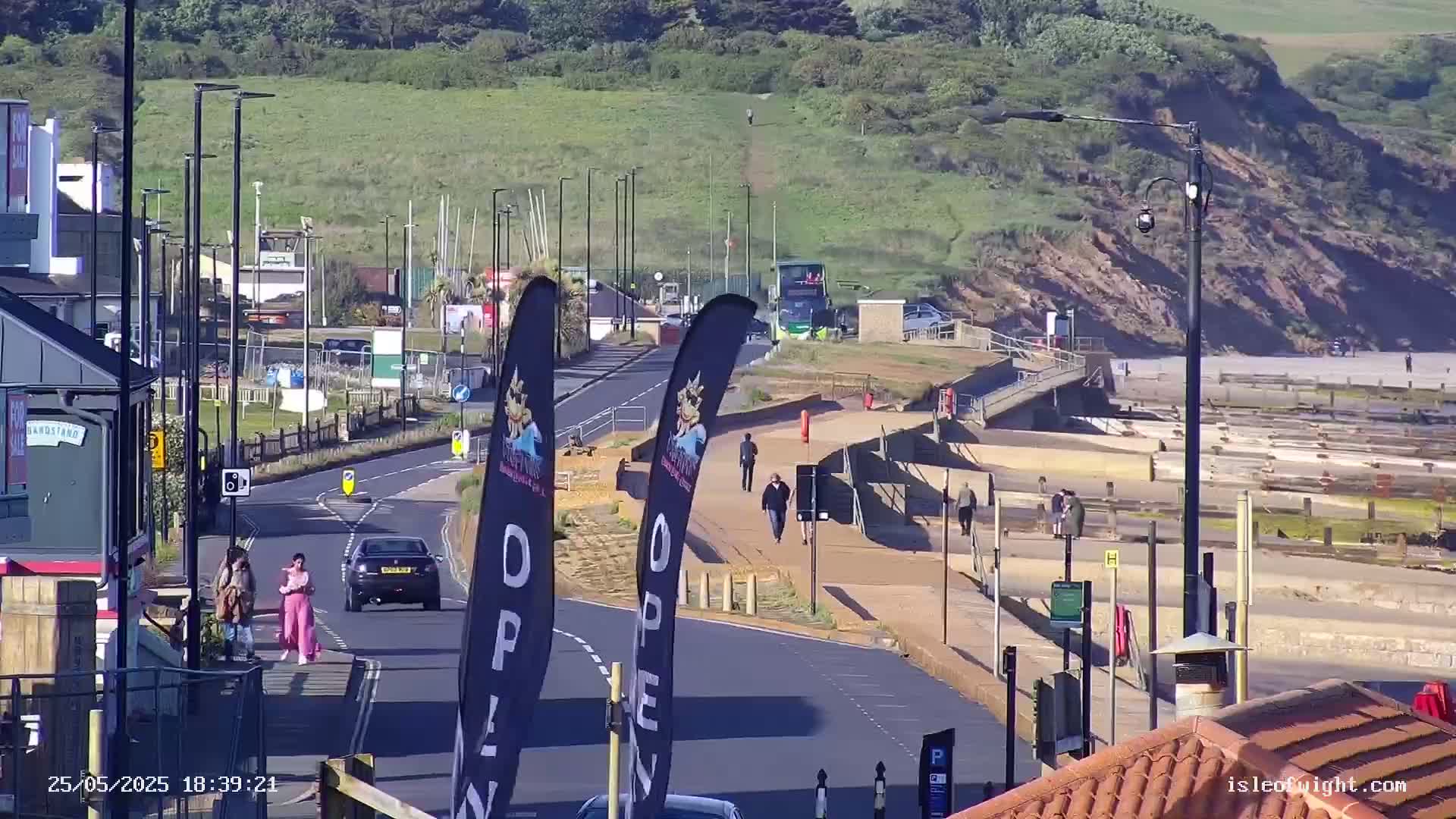 A sunny seaside town scene shows a road leading to a beach with people walking on a paved walkway, cars on the road, and a bus in the distance.