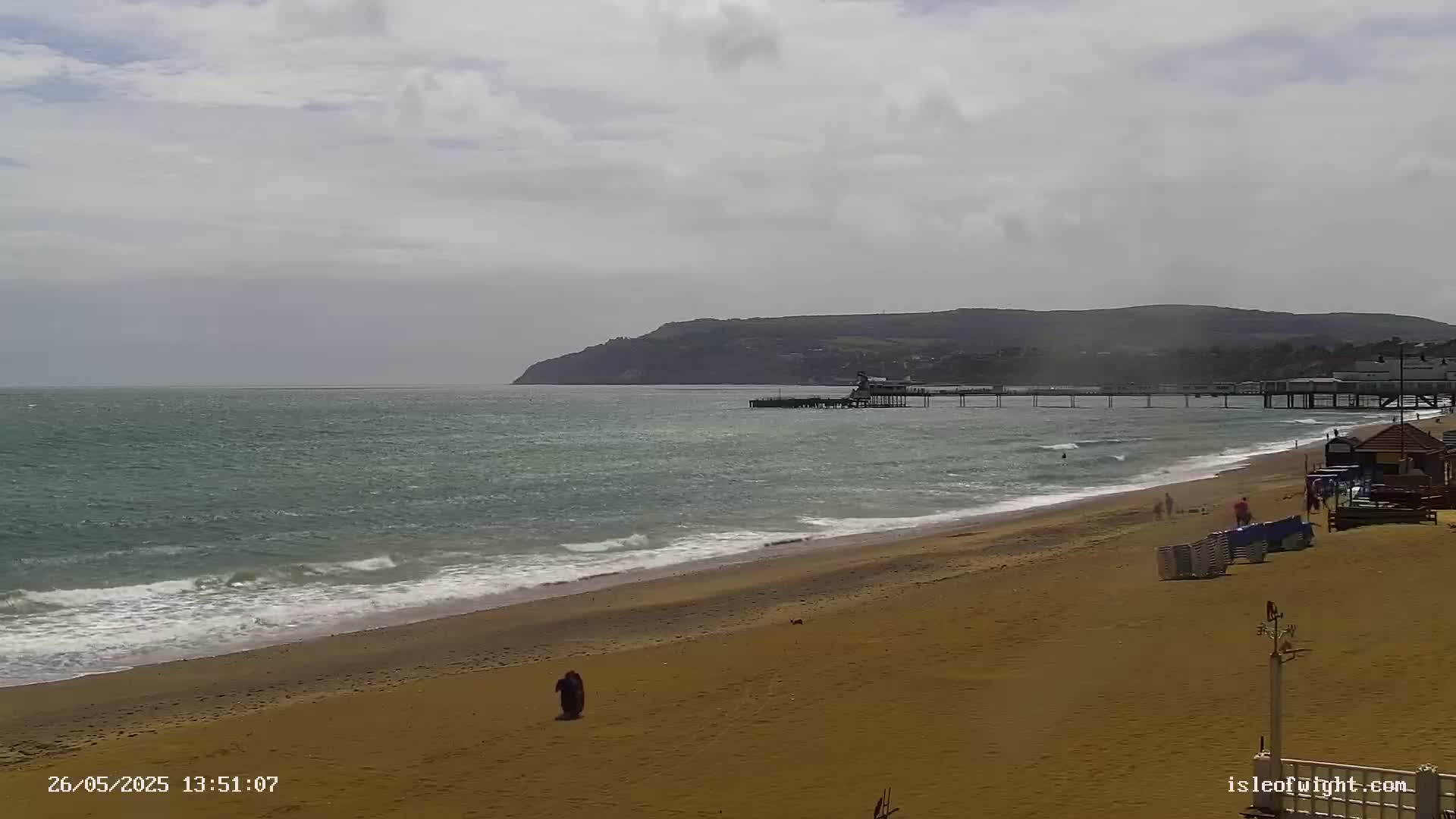 A sandy beach with a pier and a distant hill is seen under a cloudy sky, with small waves breaking on the shore.