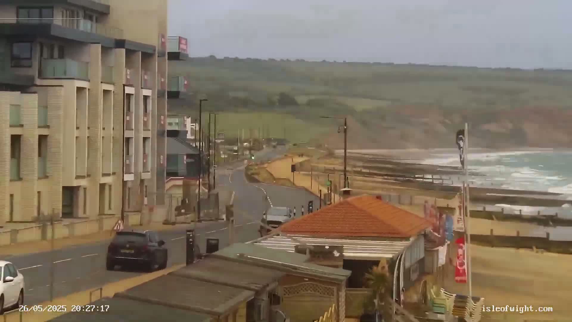 A coastal road with a modern apartment building and a small beachside building leads to a beach with moderately rough waves under an overcast sky.