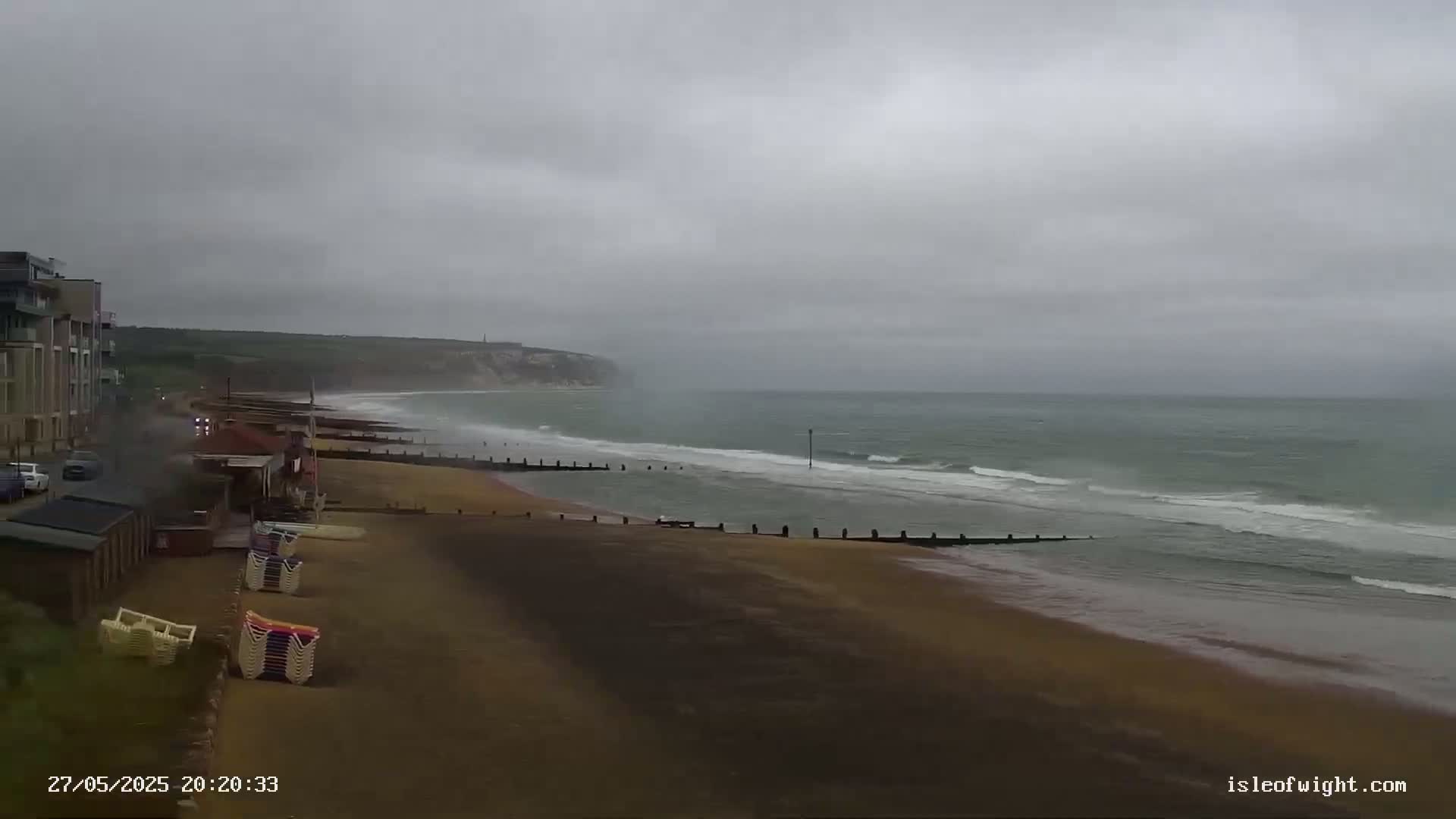 A sandy beach with stacked beach chairs and buildings is visible, next to a wavy ocean under a cloudy, overcast sky.