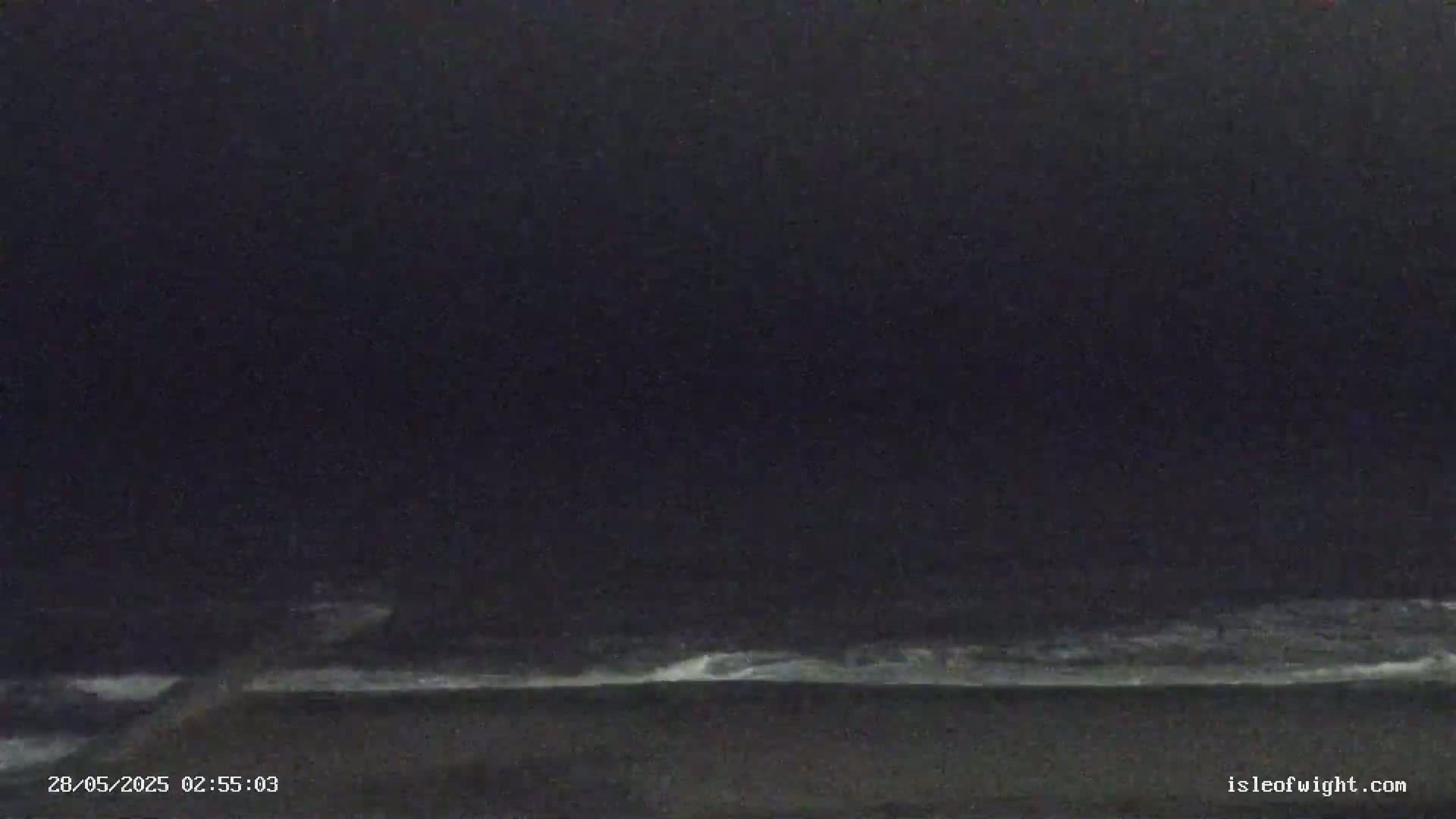 A dark nighttime scene shows a dimly lit shoreline with small waves breaking on the beach.
