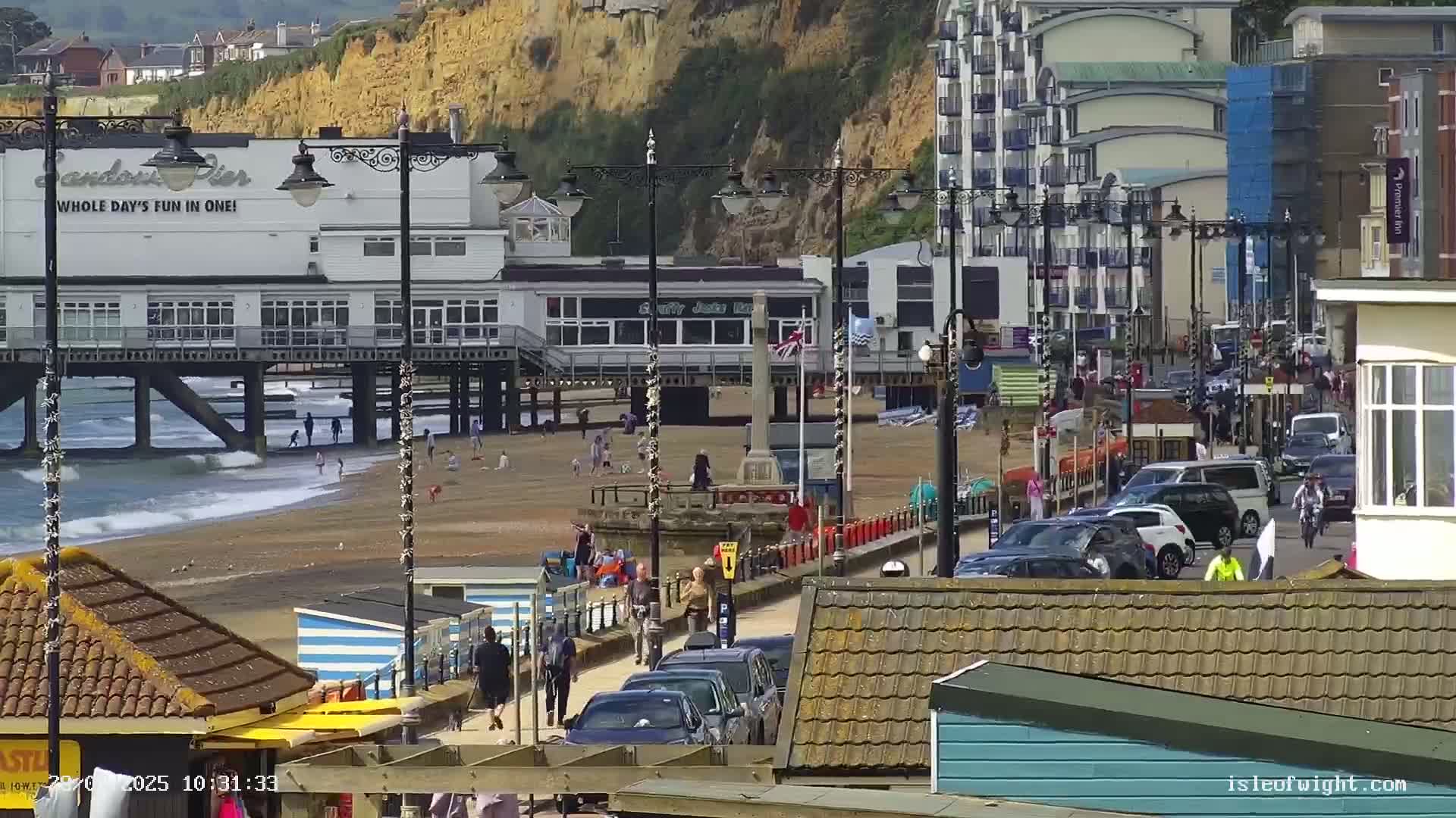 A pier with a beach and buildings behind it, under sunny skies, shows people walking and driving along a seaside promenade.