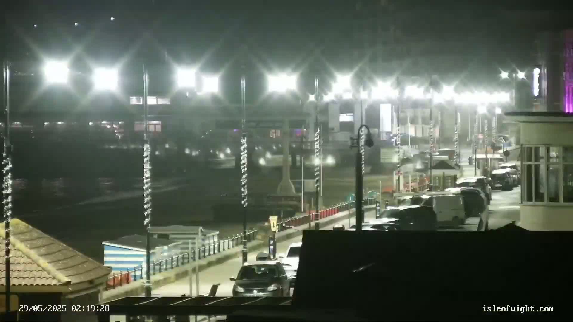 A nighttime, rain-swept seaside promenade shows several cars parked along the side of a road, lined with bright streetlights.