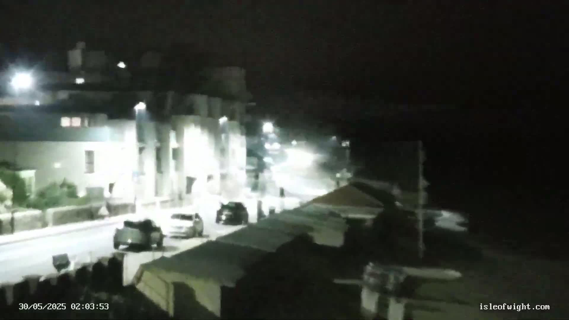 A nighttime, low-light view of a street with parked cars and buildings illuminated by streetlights.