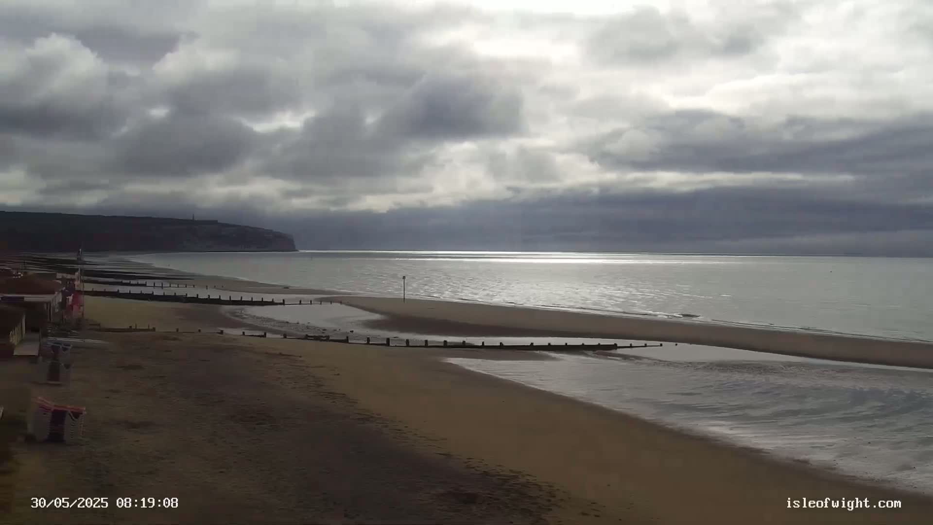 A sandy beach stretches towards a calm sea under a cloudy sky, with a distant cliff visible in the background.