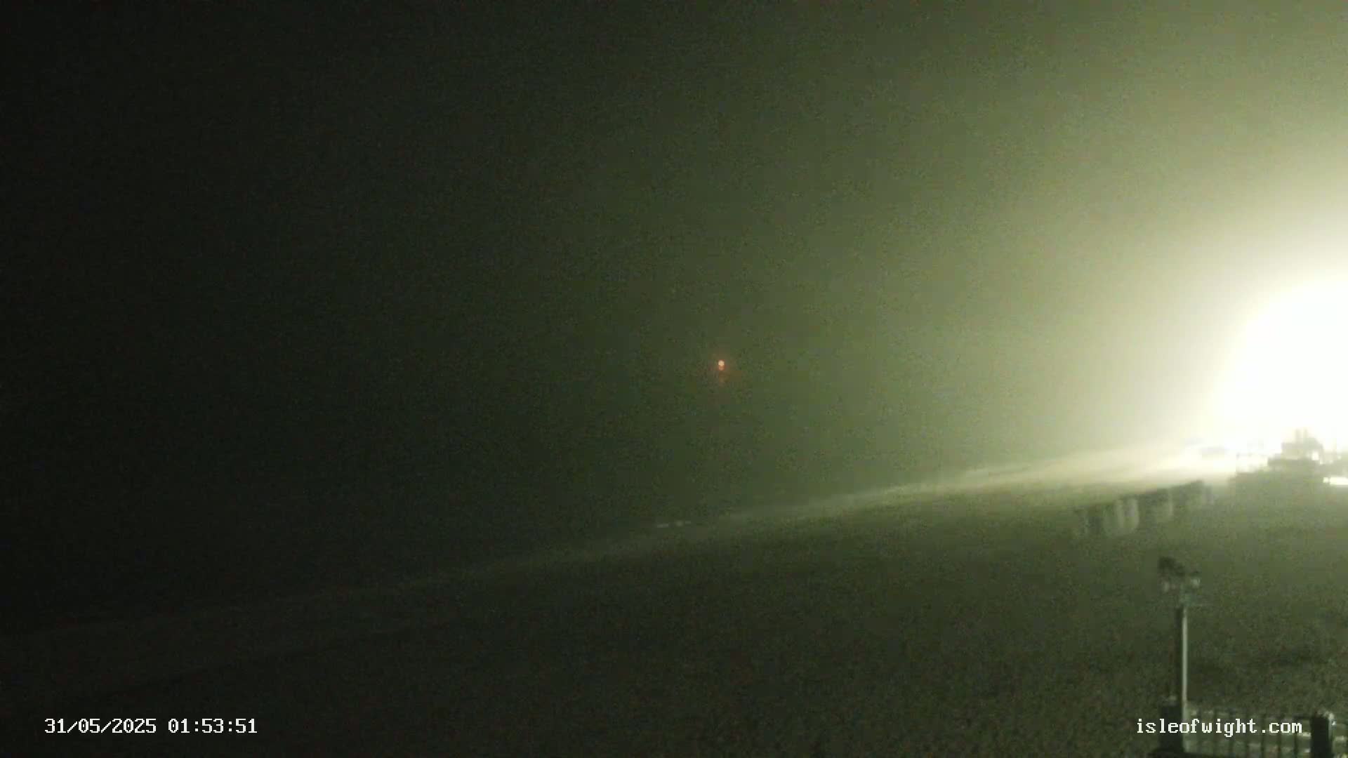 A dark beach at night is obscured by fog or mist, with a single faint light visible in the distance.