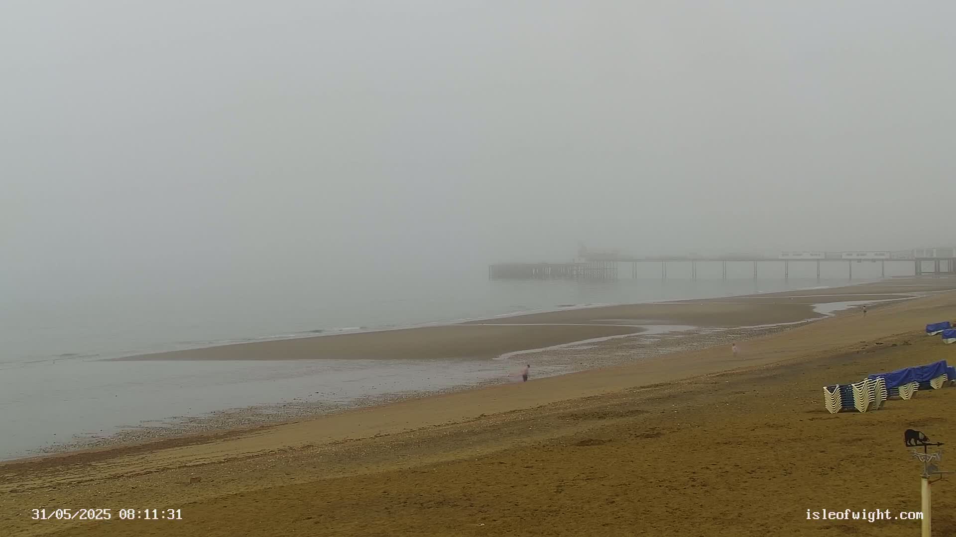 A foggy beach scene shows a pier in the distance, a sandy shore with a few people, and folded beach chairs.