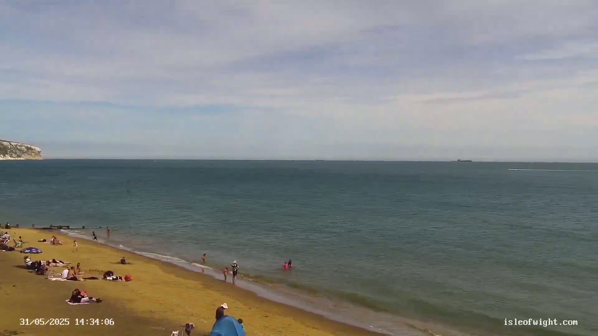 A sandy beach with people sunbathing and swimming, under a partly cloudy sky, stretches out to a calm sea with a distant white cliff in view.