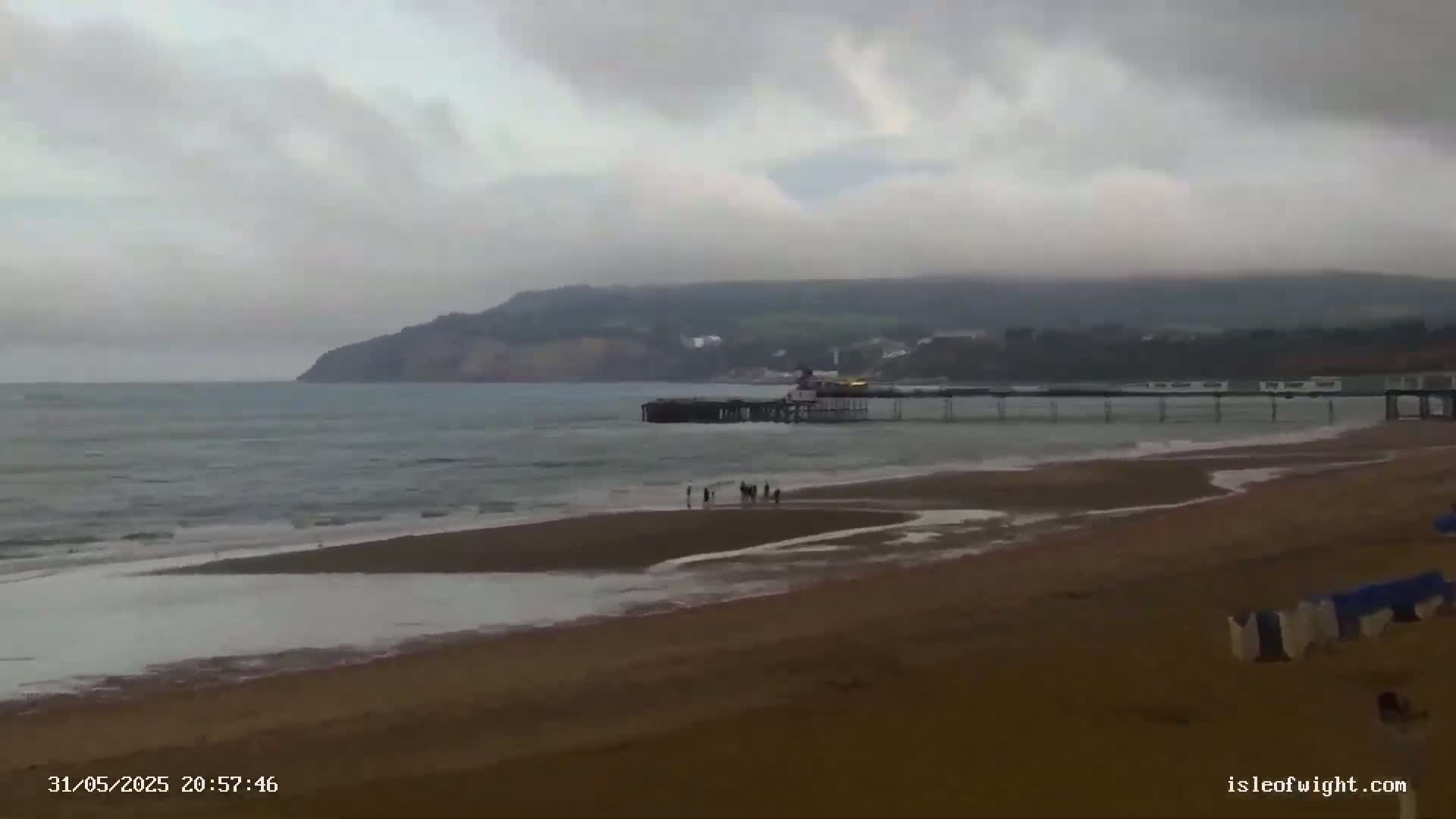 A sandy beach with a pier extending into a gray, overcast ocean, with a group of people standing near the water's edge, and a cliff visible in the distance.