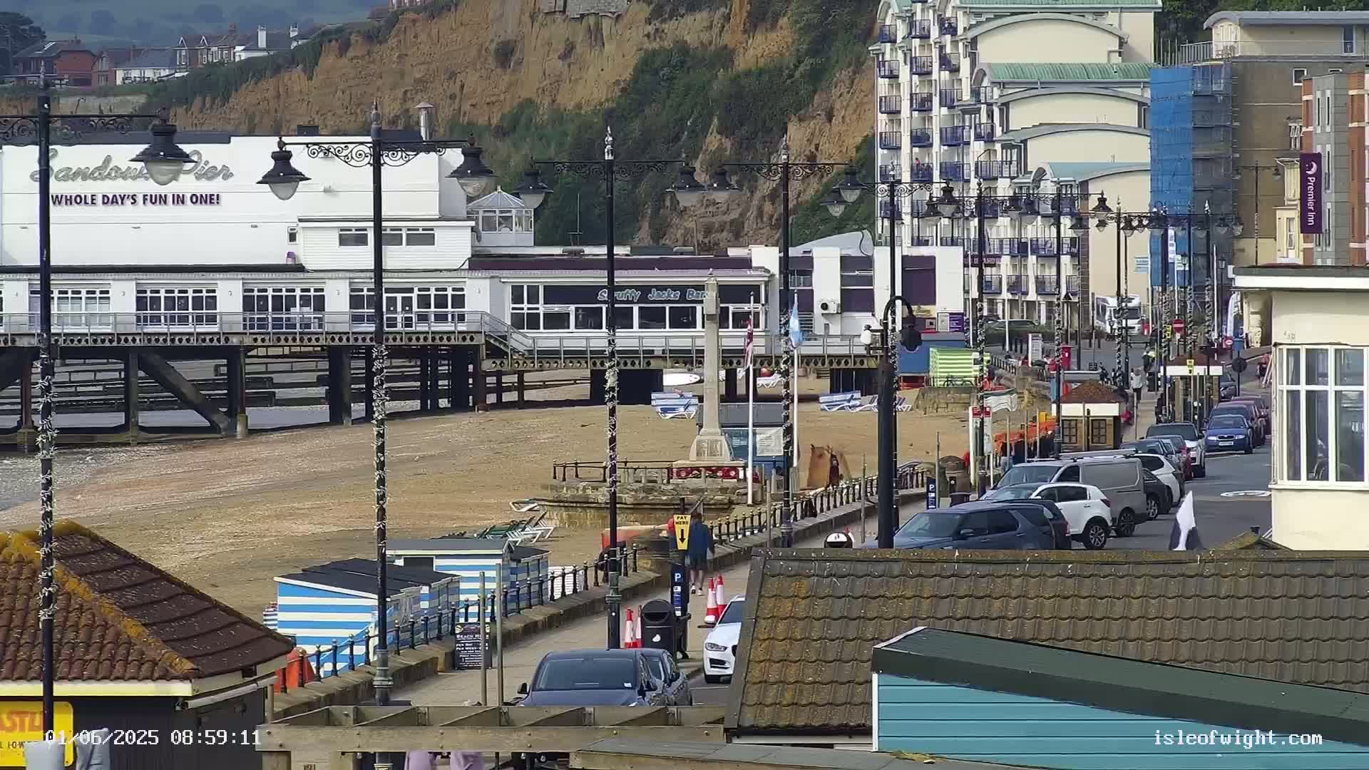 A pier with a restaurant and shops overlooks a sandy beach with parked cars and buildings on a sunny day.