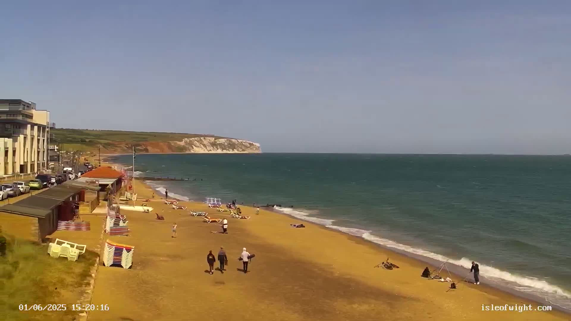 A sandy beach with a few people, beach chairs, and small buildings stretches along a calm, blue-green ocean under a clear, bright sky, with a cliff visible in the distance.