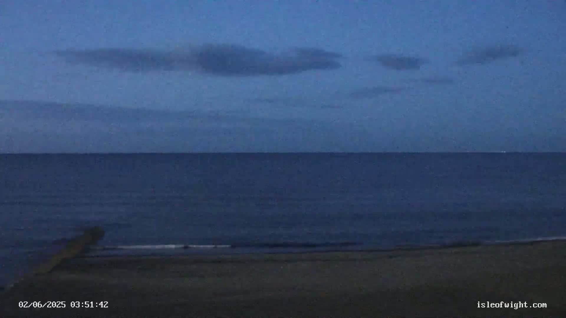 A dark blue sea meets a dark beach under a twilight sky with a few clouds.