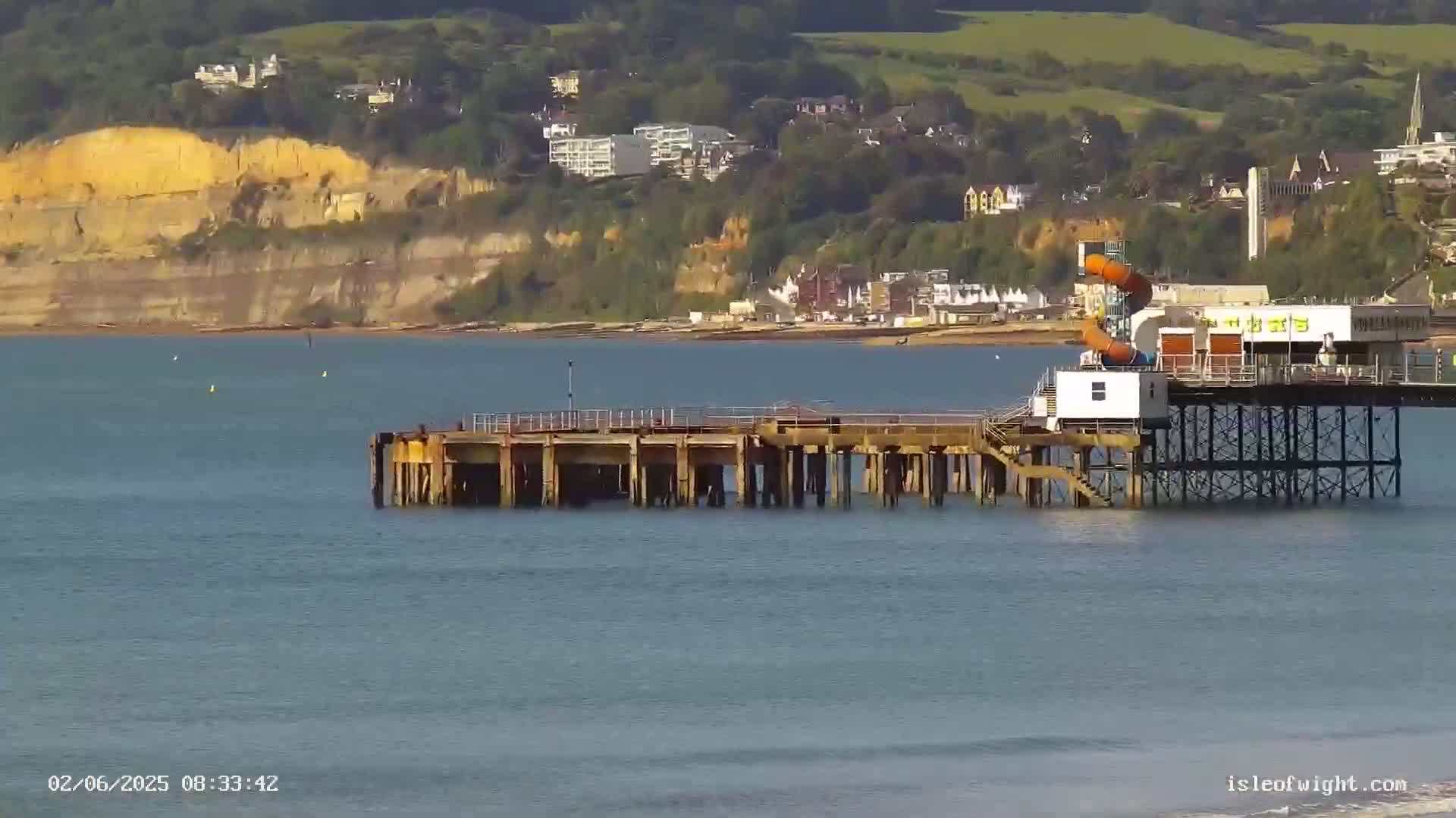 A partially dilapidated pier extends into a calm body of water, with a section featuring a water slide and small building attached to a longer pier in the background, all situated in front of a coastal town nestled against cliffs.