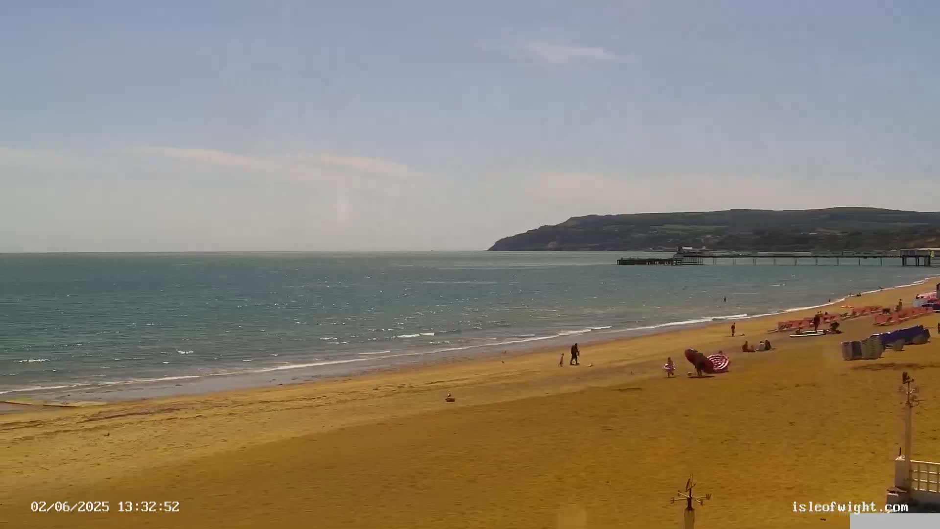 A sandy beach with a few people on it stretches to a calm sea, beyond which lies a pier and a distant hillside under a mostly sunny sky.