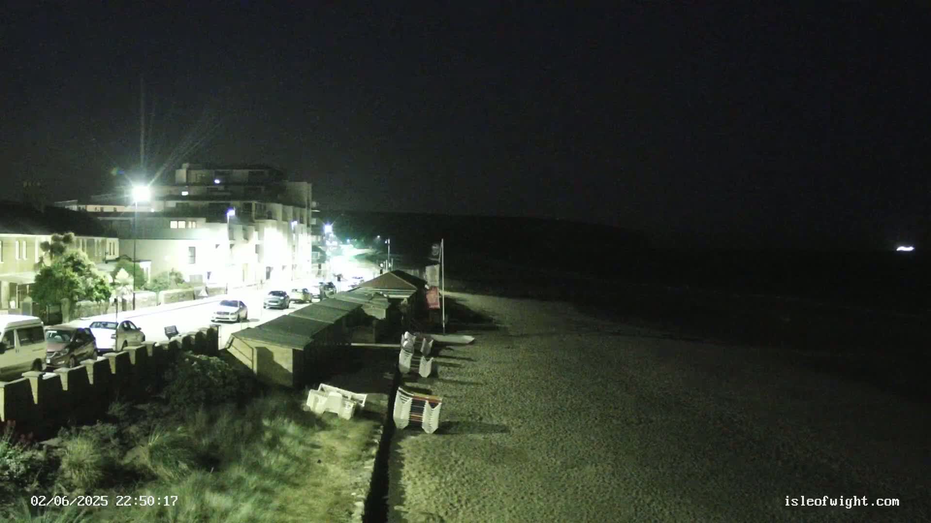 A nighttime view shows a dark beach with folded beach chairs next to a street lined with buildings and parked cars under artificial light.