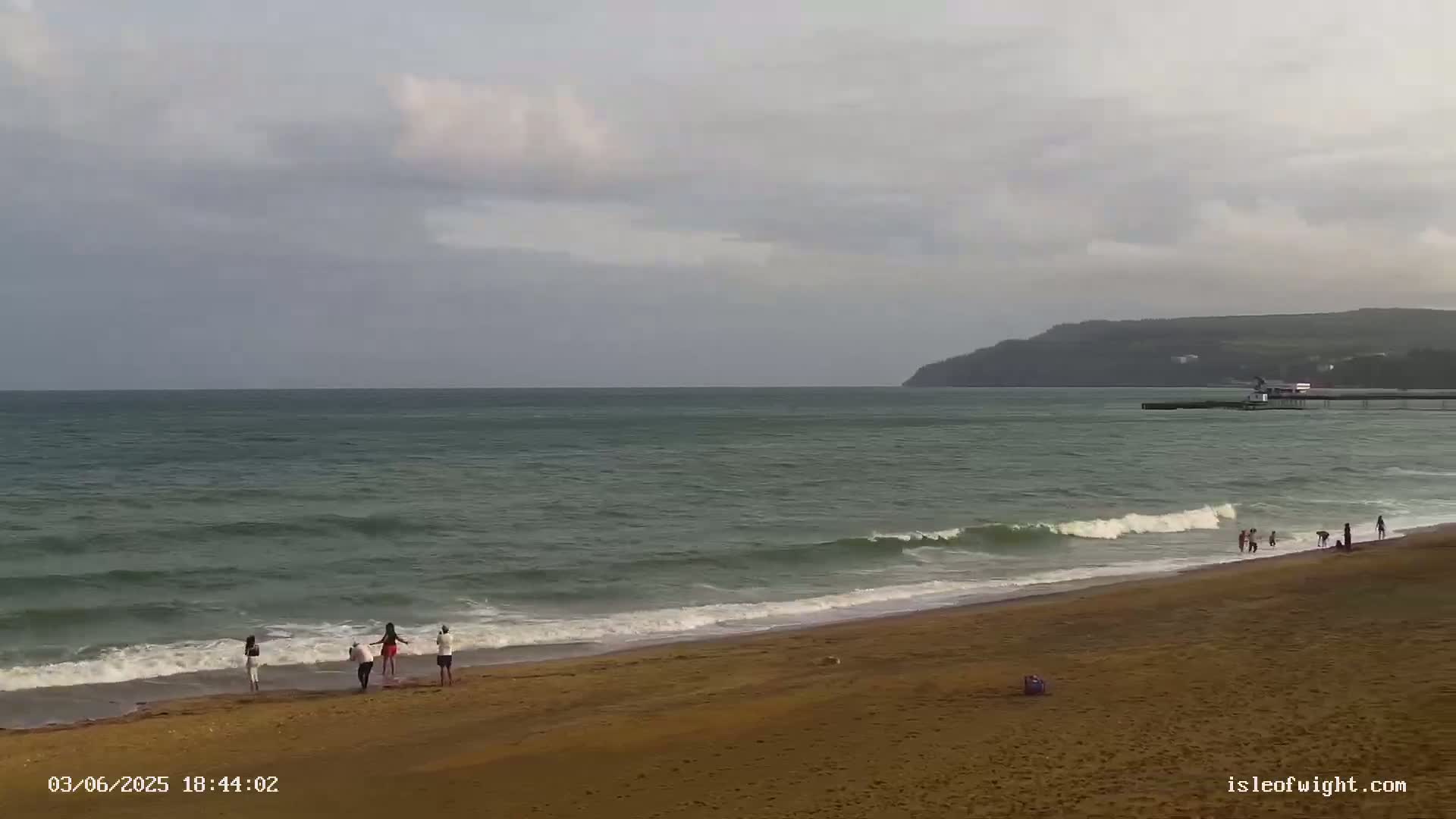 A sandy beach with several people on it is shown under a mostly cloudy sky, with gentle waves lapping the shore and a pier visible in the distance.