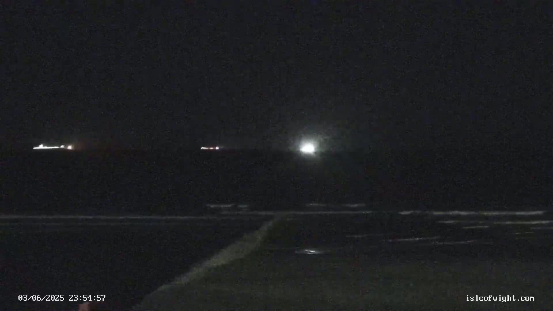 A dark nighttime beach scene shows a bright light in the distance over a gently lapping ocean.