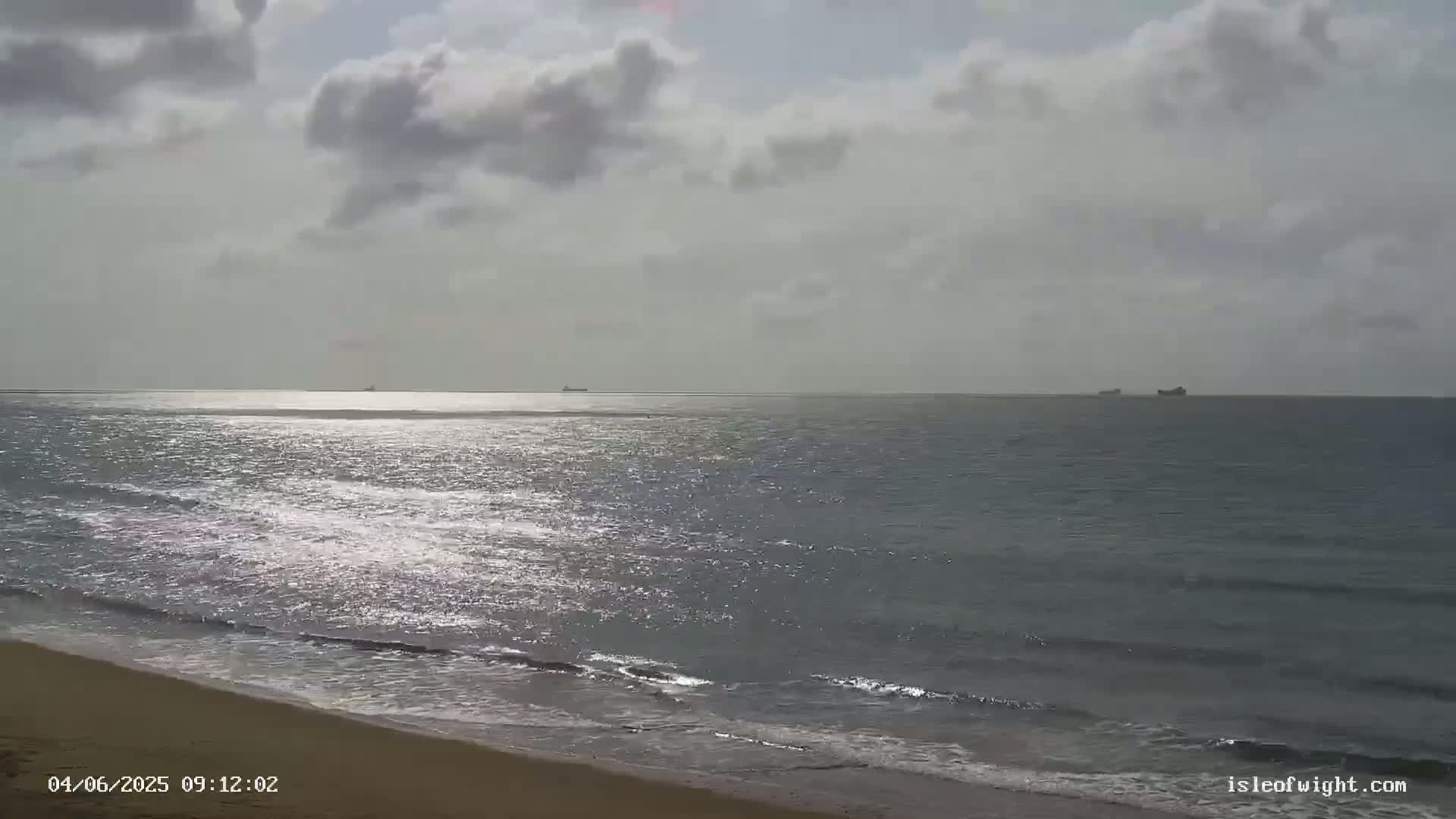 A sandy beach meets a calm, slightly shimmering sea under a mostly cloudy sky, with a few distant ships visible on the horizon.