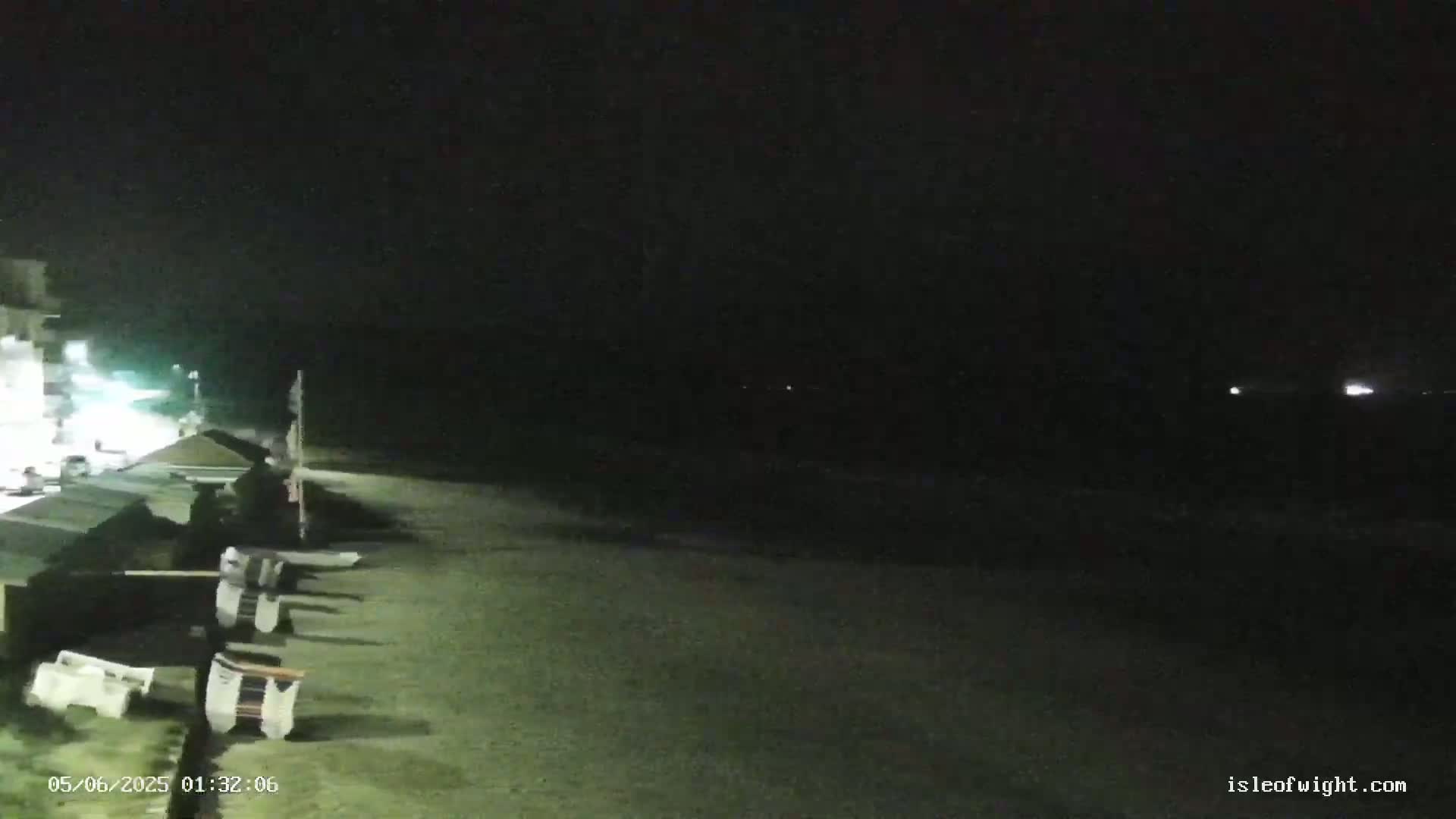 A dark, nighttime beach scene shows a row of beach chairs along a wall, with a dark ocean and faint lights in the distance.