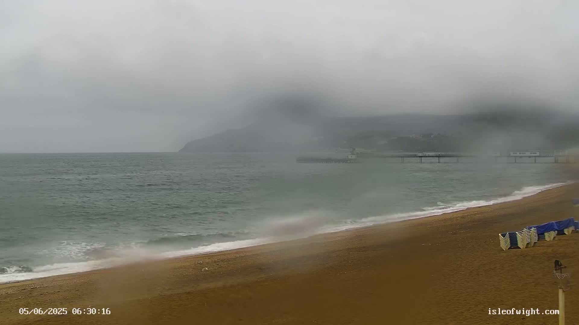 A foggy, overcast day shows a sandy beach with small waves lapping the shore, a pier in the distance, and a hill shrouded in mist in the background.