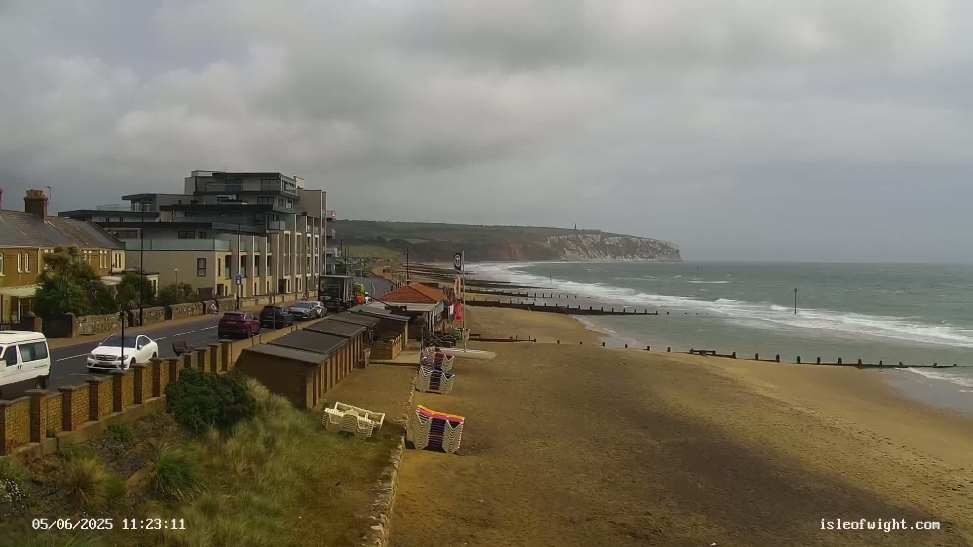 A sandy beach with small beach huts and a multi-story building is seen next to a wavy ocean under an overcast sky.