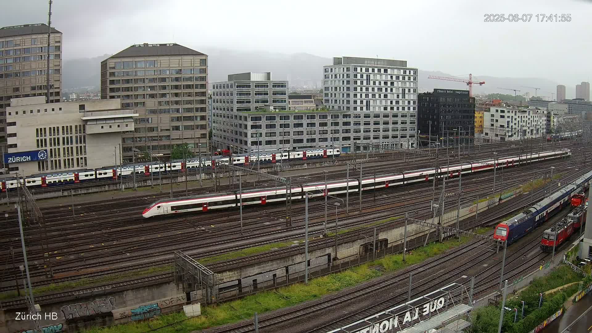 A high-angle view of a train station on a cloudy day, showing several trains on numerous tracks surrounded by modern buildings.