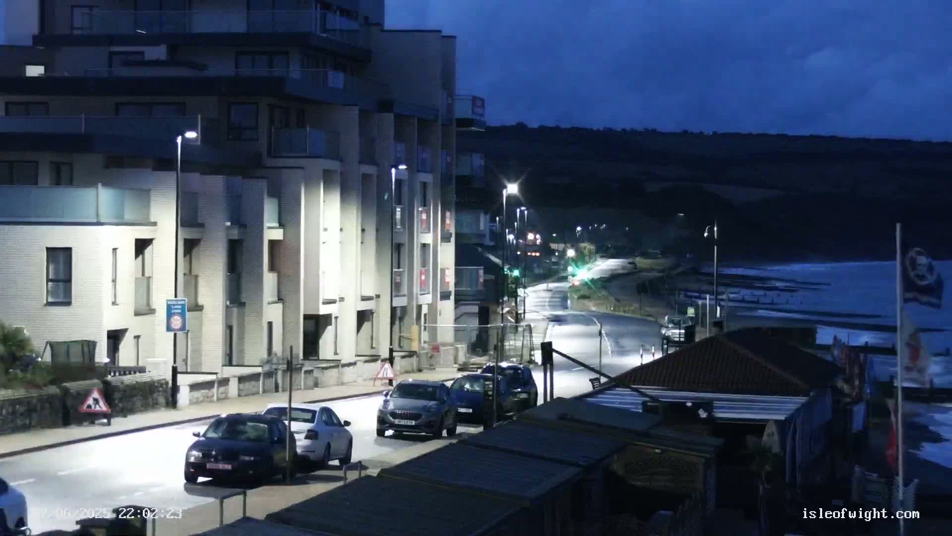 A nighttime street scene shows a multi-story apartment building, several cars on a road near the ocean, and a dark, wavy ocean under a twilight sky.